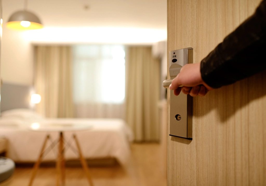 Timber guestroom door being opened by hand with the hotel room beyond